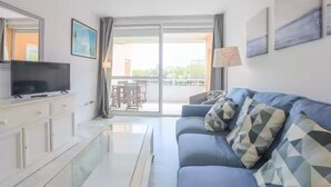 A modern living room with a flat-screen TV, a sofa with patterned cushions, a glass coffee table, and a sliding glass door leading to an outdoor area. - Las Americas - Apartment In Isla Cristina (, Huelva)