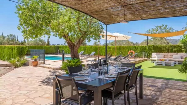 An outdoor patio with a dining table set for six, surrounded by chairs, under a shaded area with a pool in the background.