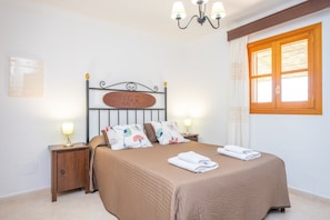 Un lit bien fait, avec une tête de lit, deux tables de chevet avec des lampes, une fenêtre avec des volets en bois et un luminaire fixé au mur.