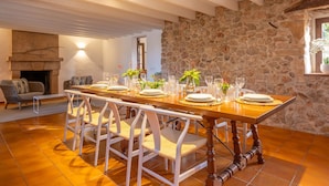 A dining room with a long wooden table set for six, surrounded by white chairs. A stone wall and a fireplace are visible in the background.