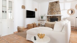 A living room with a white sofa, a stone fireplace, a flat-screen TV, and a white cabinet with glass doors. - Ca Ne Margalide - Villa With Private Pool (, Illes Balears)