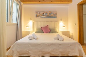A neatly made bed with a white bedspread, two pillows, and two rolled towels. There are two bedside lamps, a painting of a coastal scene on the wall, and a wooden ceiling. - Can Moreu - Villa With Private Pool In Andratx (Andratx, Illes Balears)