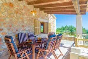 A stone wall with a wooden table and chairs, a hanging light fixture, and a view of greenery. - S'aresta Den Rumbet - Villa With Private Pool (Llucmajor, Illes Balears)