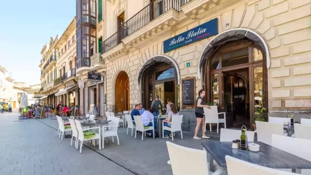 An outdoor cafe with tables and chairs, a sign for 'Bella Italia', and a building facade with arched windows and balconies. - S'aresta Den Rumbet - Villa With Private Pool (Llucmajor, Illes Balears)