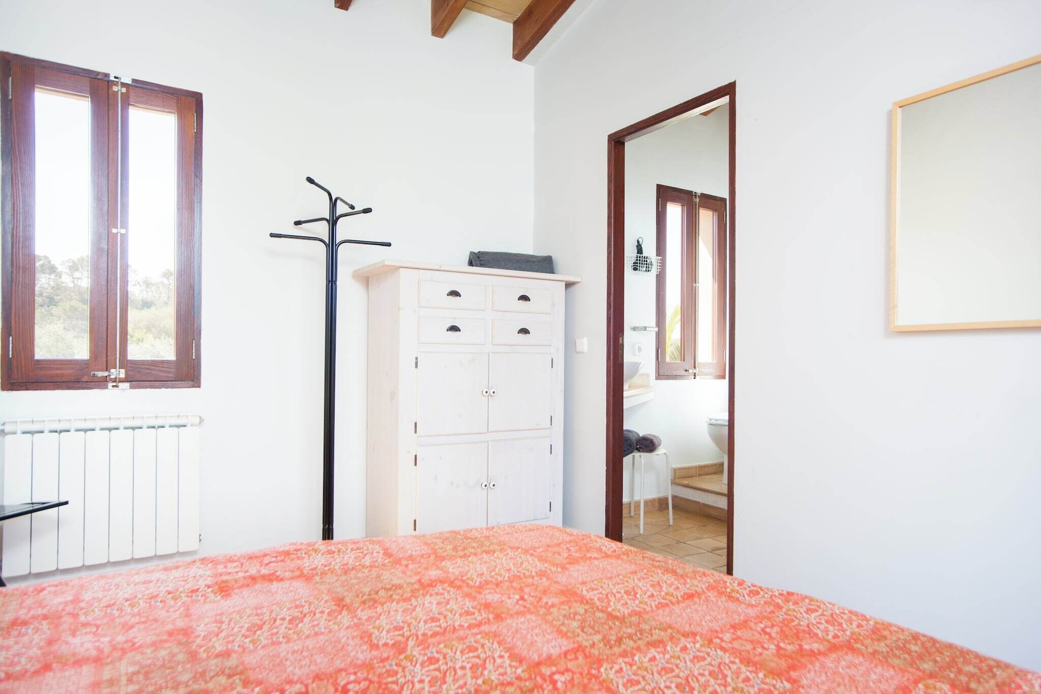 Ein Schlafzimmer mit einem Bett, einem Heizkörper, einer weißen Kommode, einem Kleiderständer und einer Tür zu einem anderen Raum.