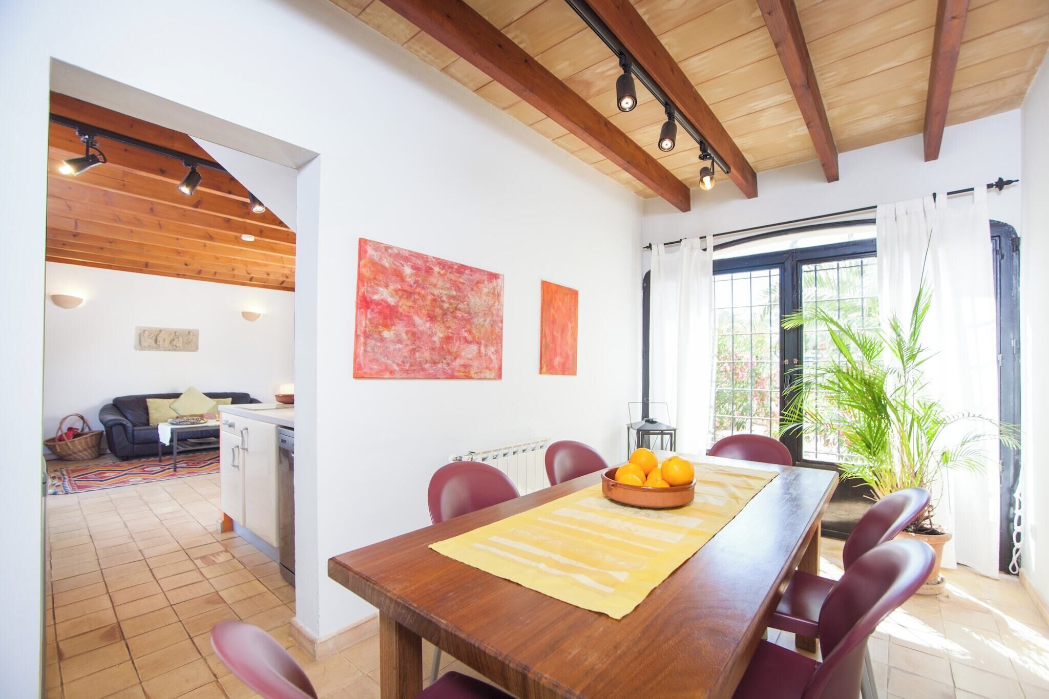A dining area with a wooden table, red chairs, and a bowl of oranges. There is a kitchen in the background with a black countertop and a white cabinet.