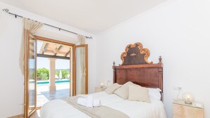 A bedroom with a wooden headboard, a bed with white linens, a glass side table with a lamp, and a view of a swimming pool through an open door. - Son Roca - Villa With Private Pool In Montuïri (Montuïri, Illes Balears)
