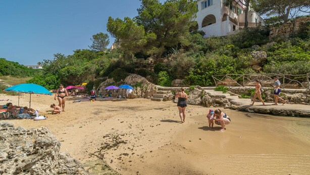Una playa con gente tomando el sol y caminando, una costa rocosa y un edificio al fondo.