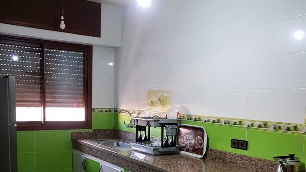 Une cuisine avec des meubles de couleur verte, un plan de travail en granit, une cuisinière et une fenêtre équipée de stores.