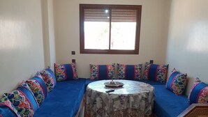 Une pièce comprenant un long canapé bleu, une table ronde recouverte d’une nappe à motifs et une fenêtre avec des stores.