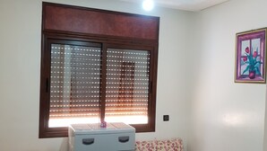 Une chambre avec deux lits, une commode, une fenêtre avec des stores, un luminaire au plafond et un tableau accroché au mur.