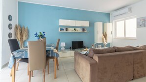 A modern living room with a blue accent wall, a brown sofa, a dining table with chairs, a flat-screen TV, and a bookshelf. - Amazing and Spacious Apartmet wSide Sea View (San Pawl il-Baħar)