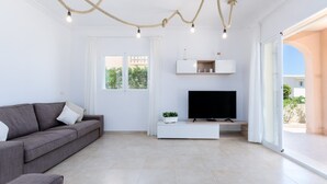 A modern living room with a grey sofa, a flat-screen TV on a white stand, and a large sliding glass door leading to an outdoor area. - Dones D'aigua 11 - Villa With Private Pool (S'estanyol (colònia De Sant Pere), Illes Balears)
