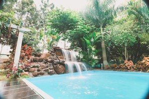 A swimming pool with a waterfall feature surrounded by lush greenery and rocks. - Emerald - 2BR Pool Villa Near Airport (Bengaluru)