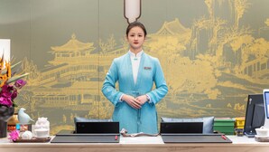 A reception desk with a traditional Chinese mural in the background, a blue uniformed staff member, and various items on the desk including a laptop, a tablet, and a small decorative vase. - The Serene House (Hangzhou)