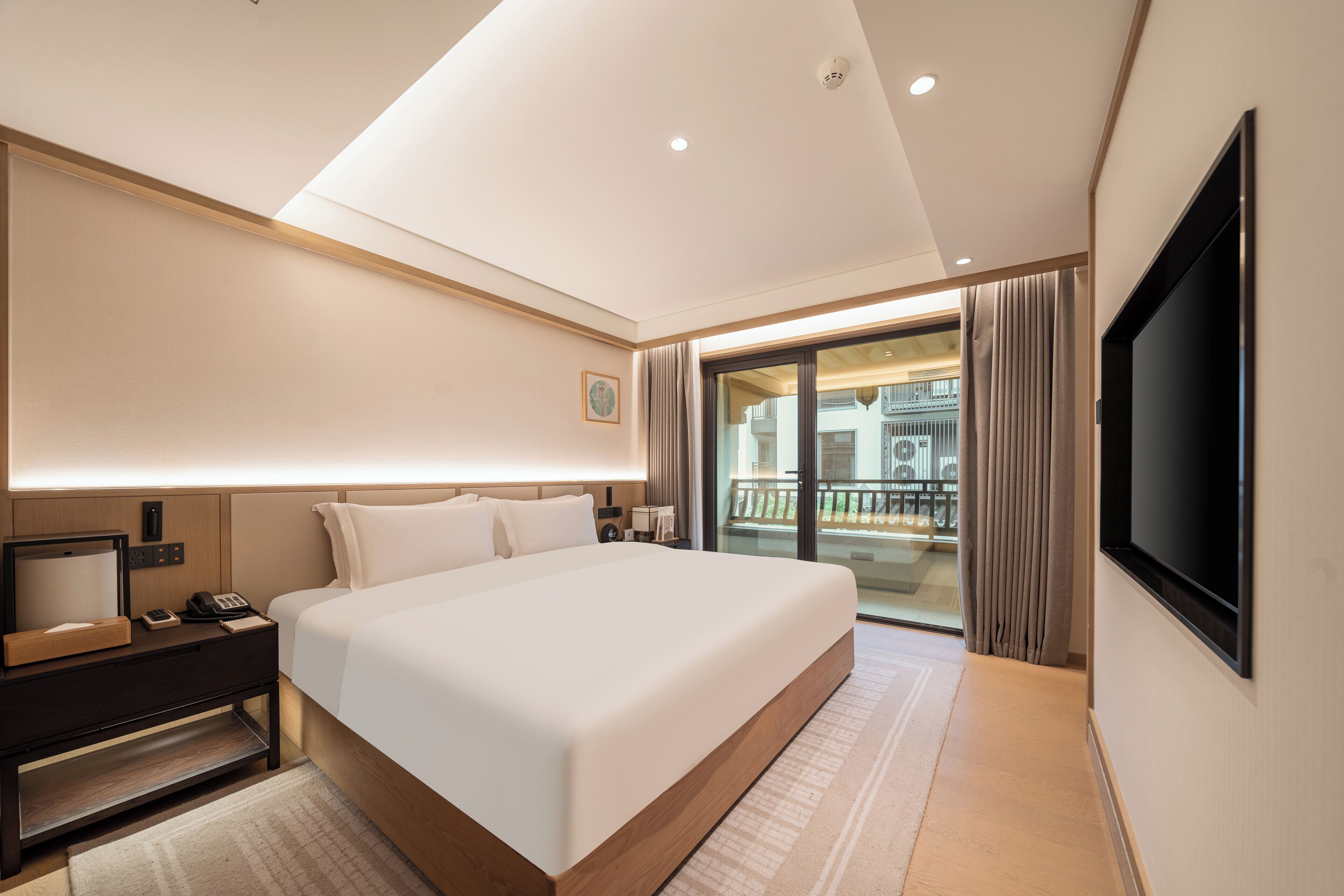 A modern hotel room with a large bed, a flat-screen TV, a bedside table with a lamp, and a view of the outside balcony.