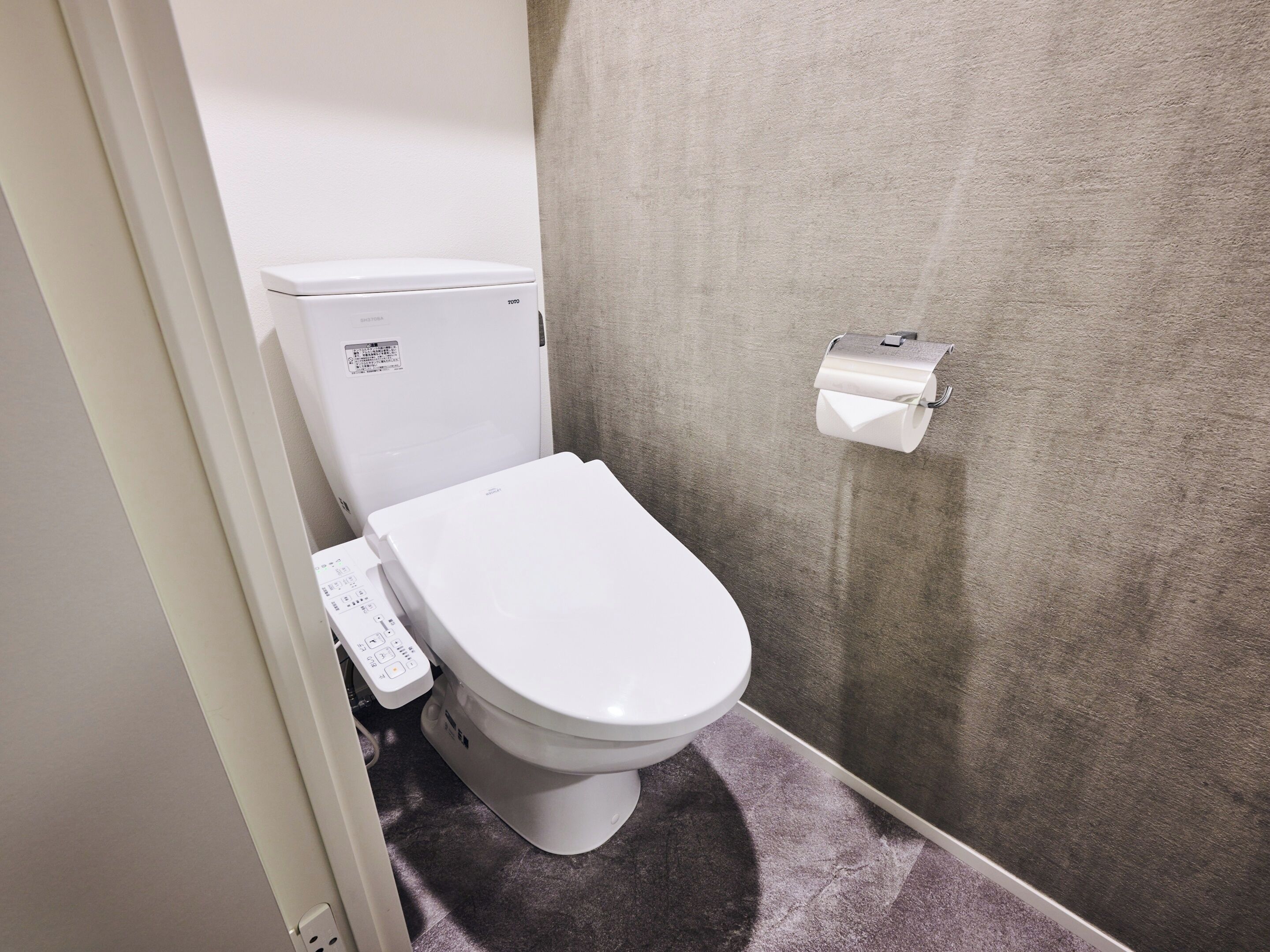 A small bathroom with a toilet, a toilet paper holder, and a control panel.
