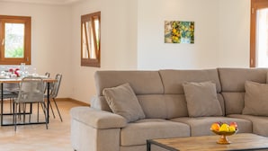 Een woonkamer met een beige bank, een houten salontafel met een fruitschaal, en een eethoek met een tafel en stoelen.
