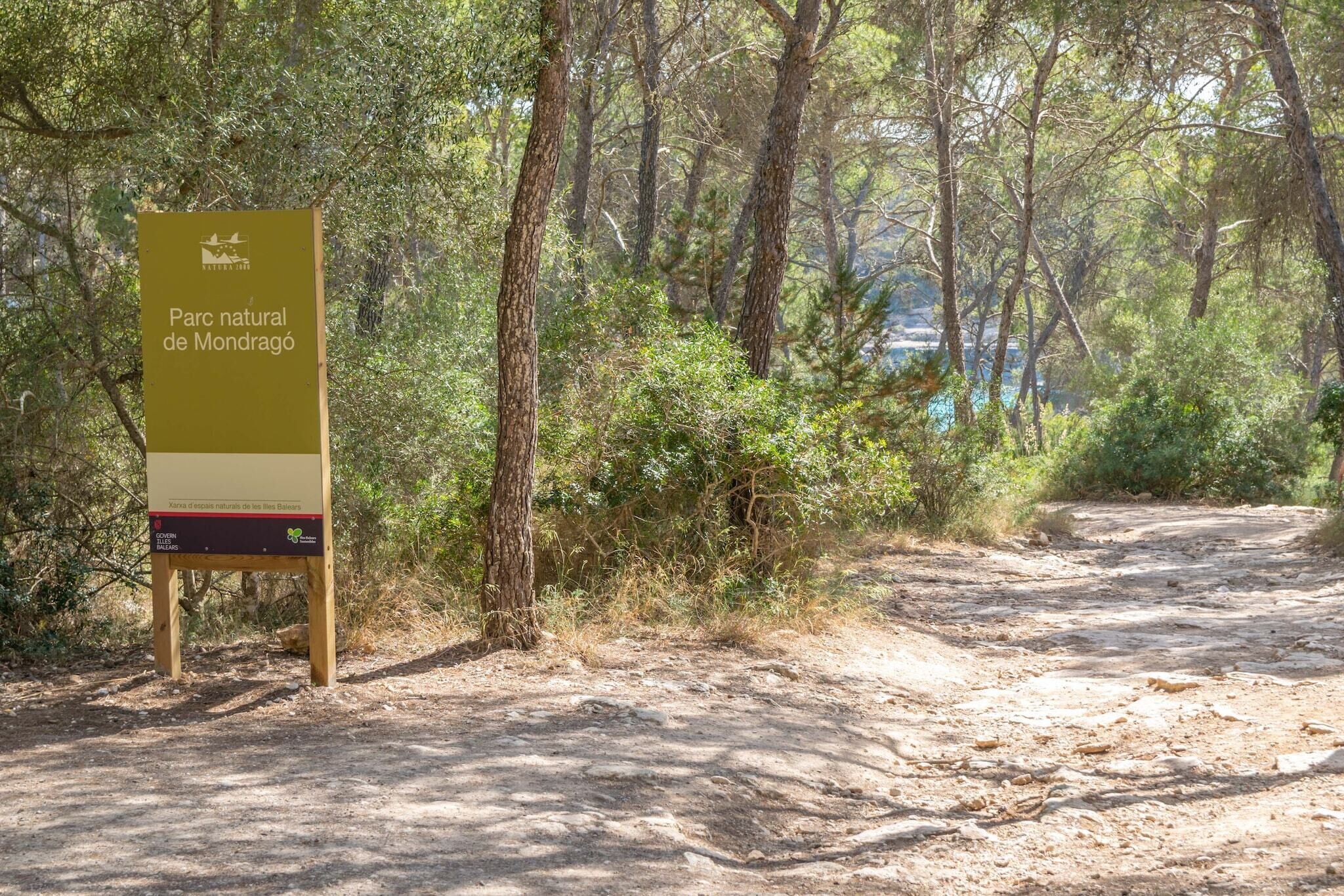 A sign for Parc natural de Mondragó with a dirt path leading through a wooded area.
