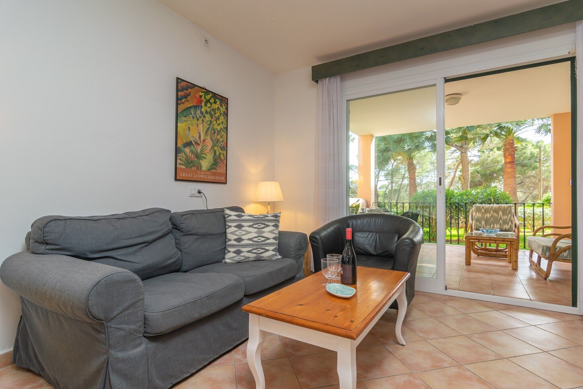 A living room with a grey sofa, a wooden coffee table, a black leather chair, and a glass door leading to an outdoor balcony with a view of palm trees and a garden.