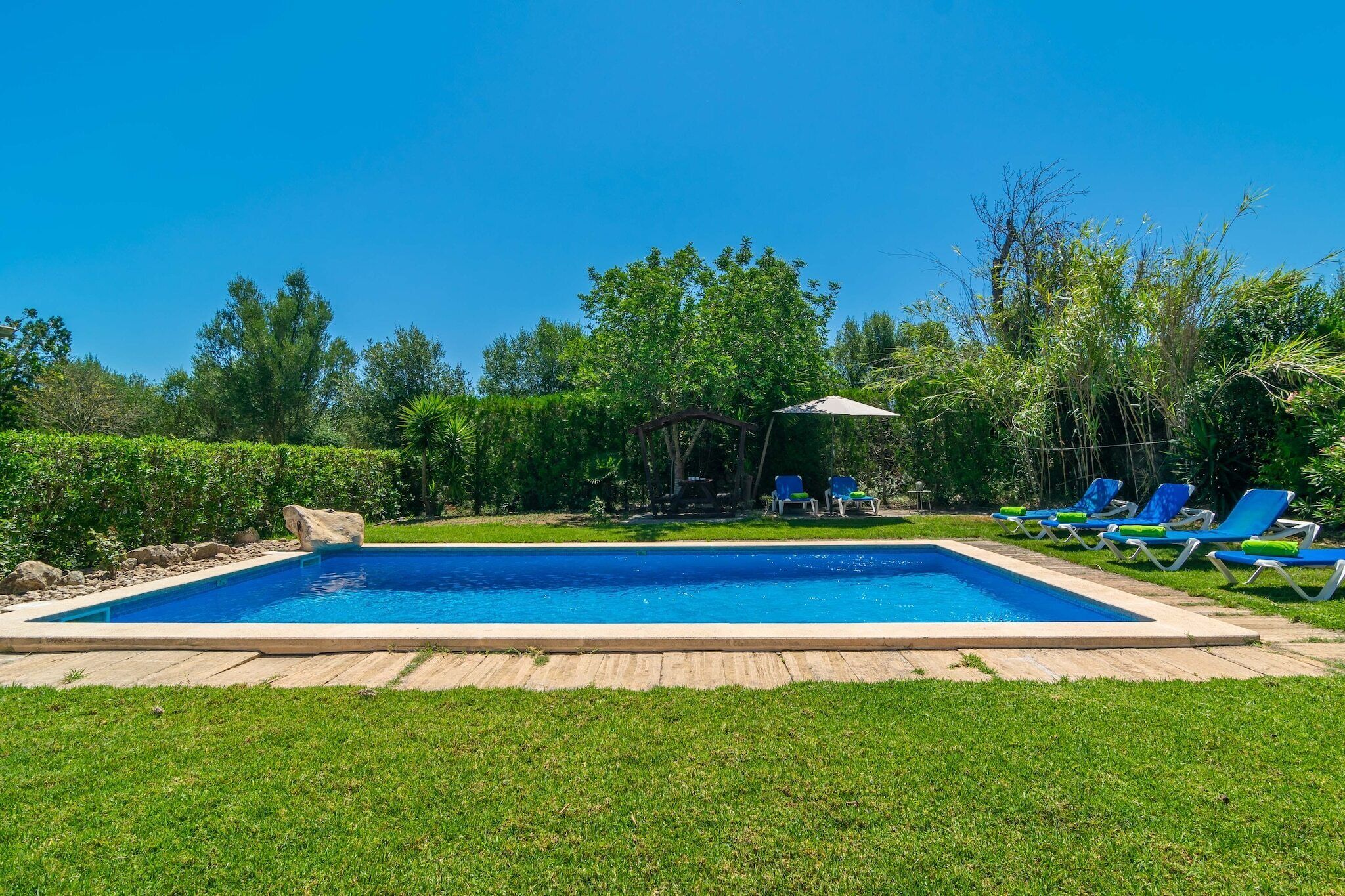 A rectangular swimming pool with a wooden deck, surrounded by a well-maintained lawn and a row of blue lounge chairs.