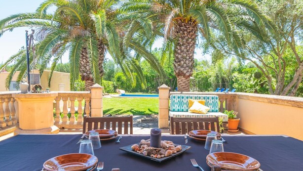 Un área de comedor al aire libre con una mesa lista para dos personas, rodeada de palmeras y vista a una piscina.