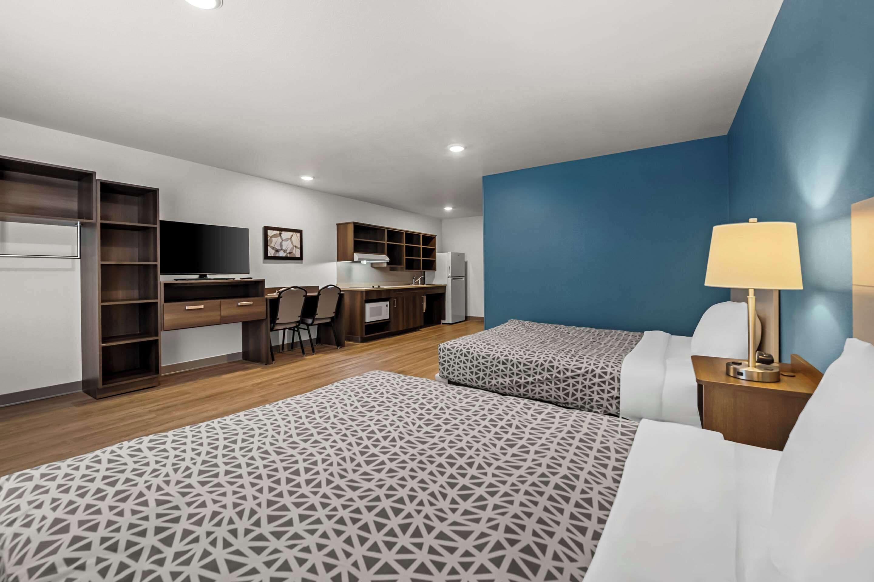 A modern hotel room with a bed, bedside tables, a desk, and a kitchenette.