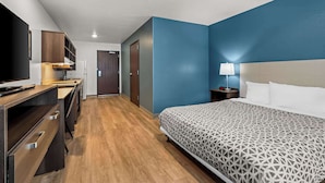 A hotel room with a bed, a desk, a television, and a nightstand with a lamp. - Woodspring Suites Loves Park Rockford (Loves Park)