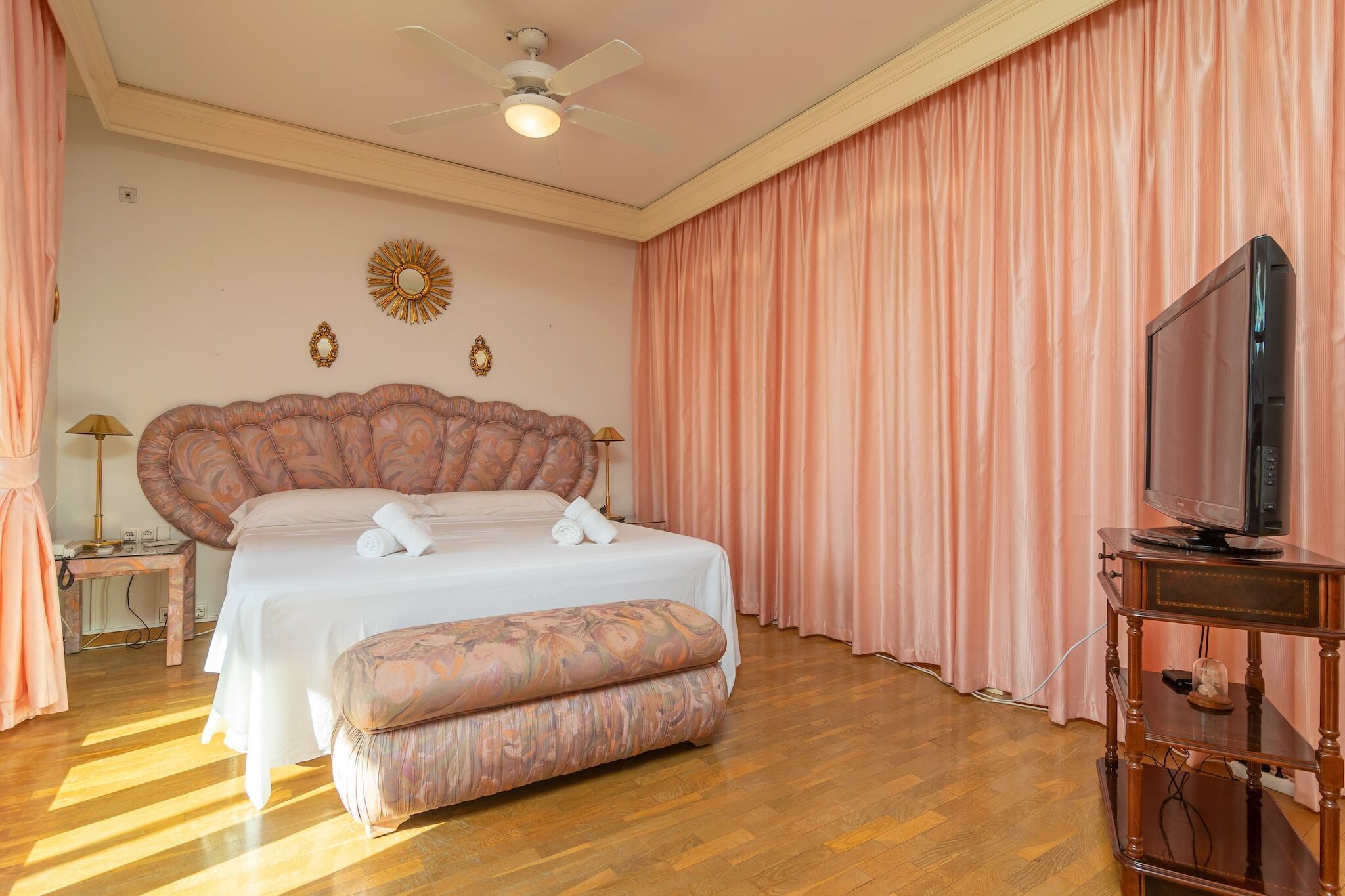 Une chambre à coucher avec un grand lit, un ventilateur de plafond, un téléviseur posé sur un meuble en bois et des rideaux roses.