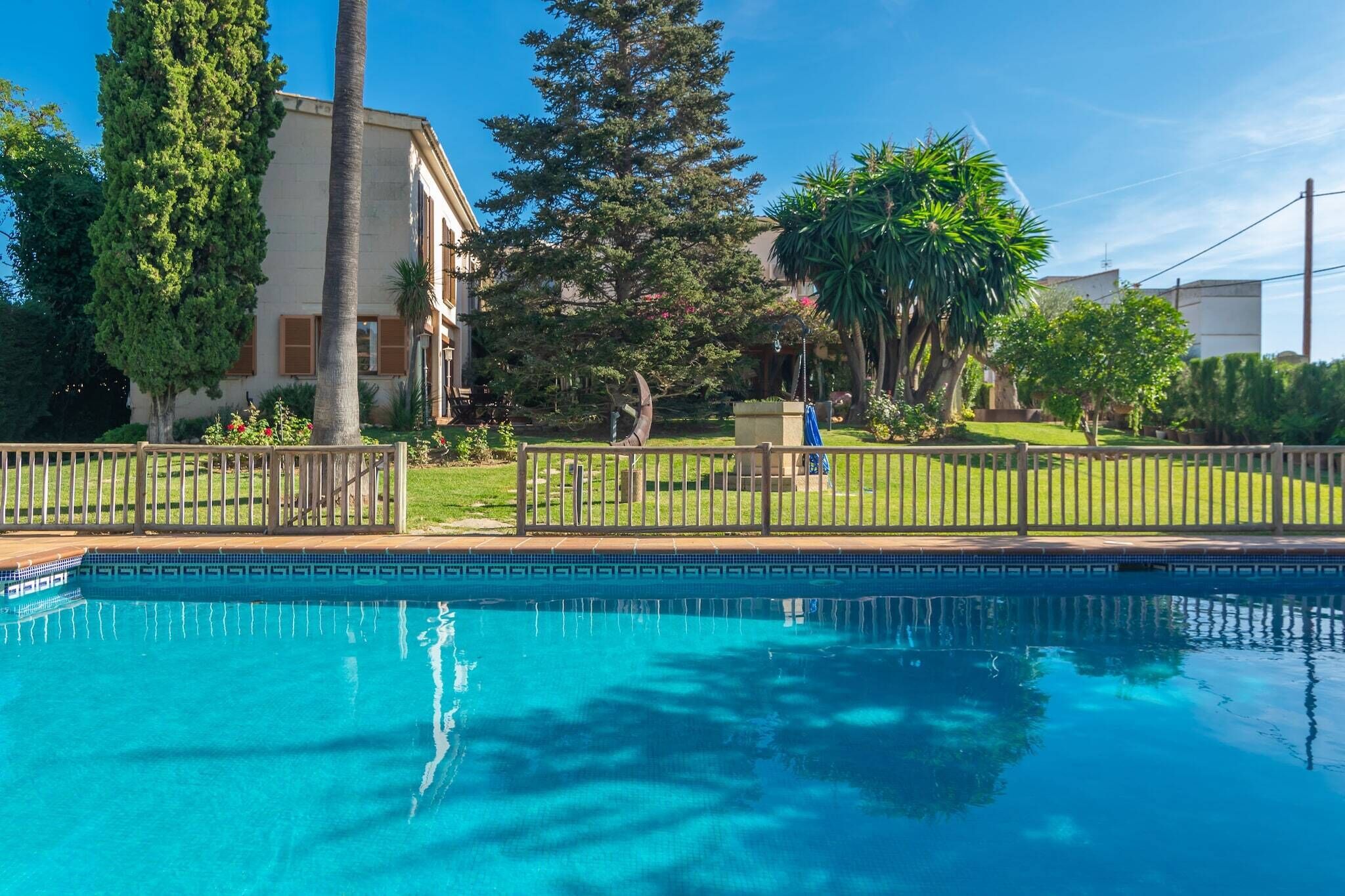 Una piscina con plataforma de madera, rodeada por una valla, y un edificio con jardín al fondo.
