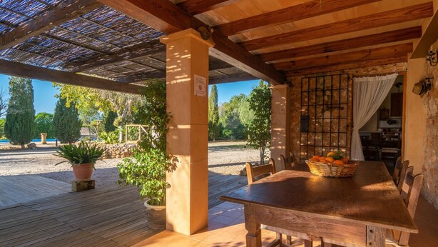 An outdoor wooden deck with a table and chairs, surrounded by potted plants and a view of a garden and trees. - Canaus - Villa With Private Pool In Porreres (Porreres, Illes Balears)