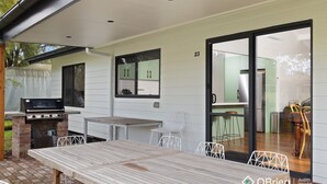 Un patio exterior con una mesa y sillas de madera, una parrilla de barbacoa y una puerta corrediza de vidrio que da a una cocina.