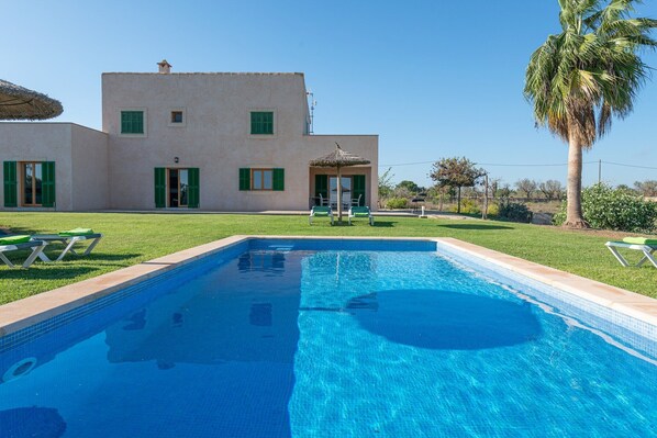 A swimming pool with a clear blue water, surrounded by a well-maintained lawn and a building with green shutters.