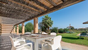 Une terrasse extérieure avec une table dressée pour le repas, entourée de chaises blanches, et protégée par une structure de toit en chaume.