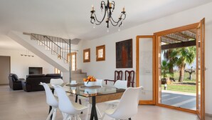 Un vaste espace de vie comprenant une table à manger, des chaises blanches, un escalier et une grande fenêtre donnant sur des palmiers.