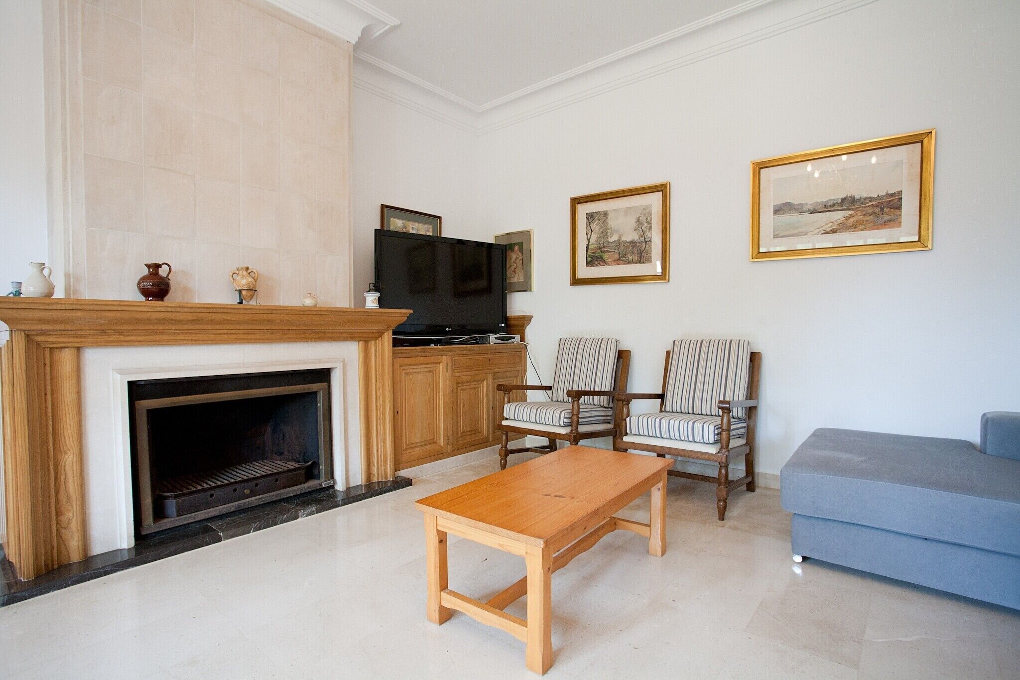 Un salon avec une cheminée, un téléviseur, deux fauteuils à rayures, un canapé bleu et des tableaux encadrés accrochés au mur.