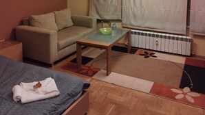 Habitació d'hotel amb sofà, taula petita amb un bol i ampolla d'aigua, radiador i llit amb llençol estampat.