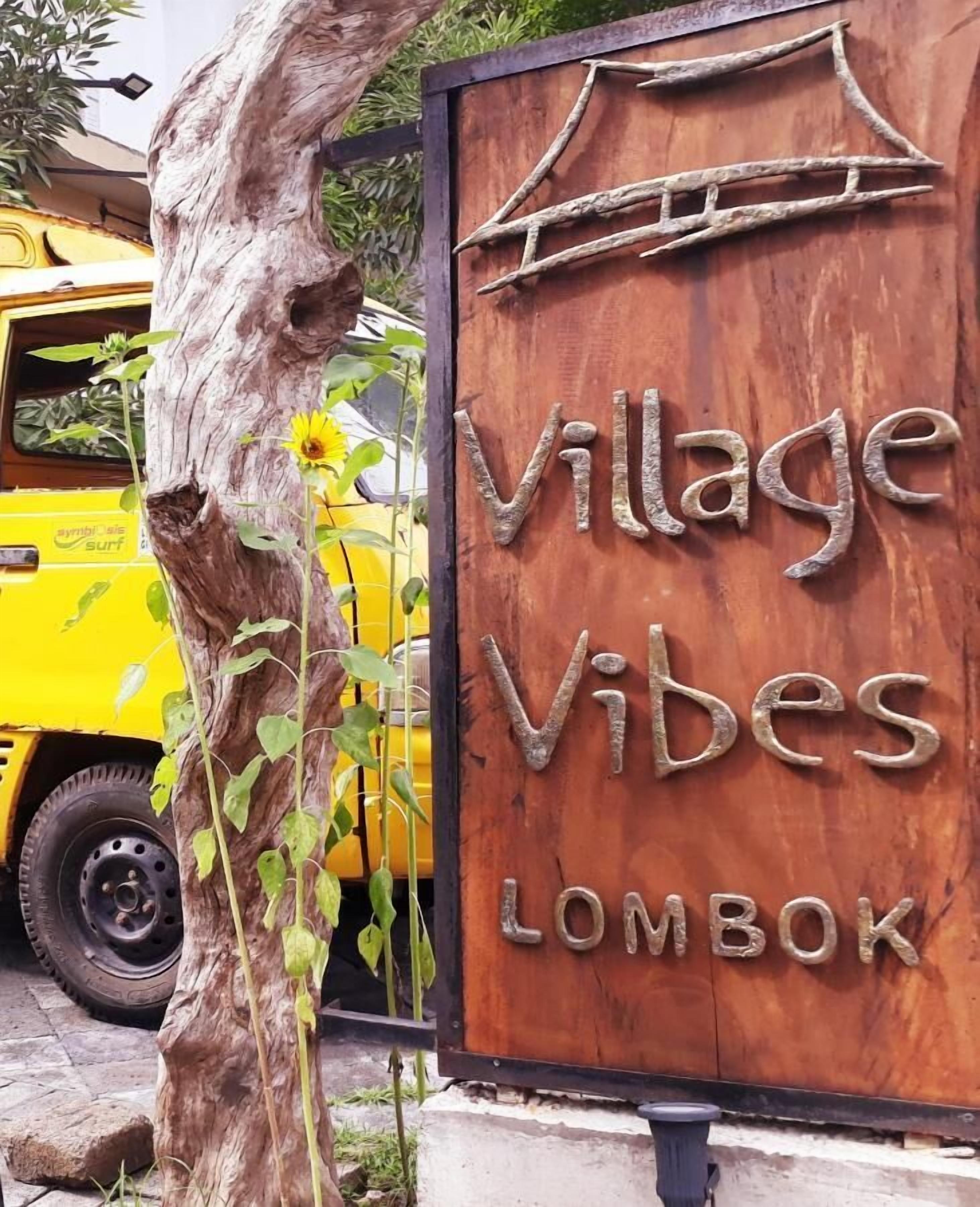 A wooden sign with the text "Village Vibes LOMBOK" and a tree with green leaves in the foreground.