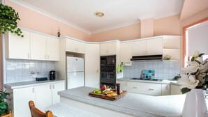 Une cuisine moderne avec des meubles blancs, un îlot central agrémenté de fruits et de pâtisseries, un réfrigérateur, un four à micro-ondes et un four traditionnel.