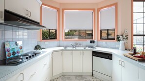 Une cuisine avec des meubles blancs, une cuisinière à gaz, un évier et une fenêtre avec des stores.