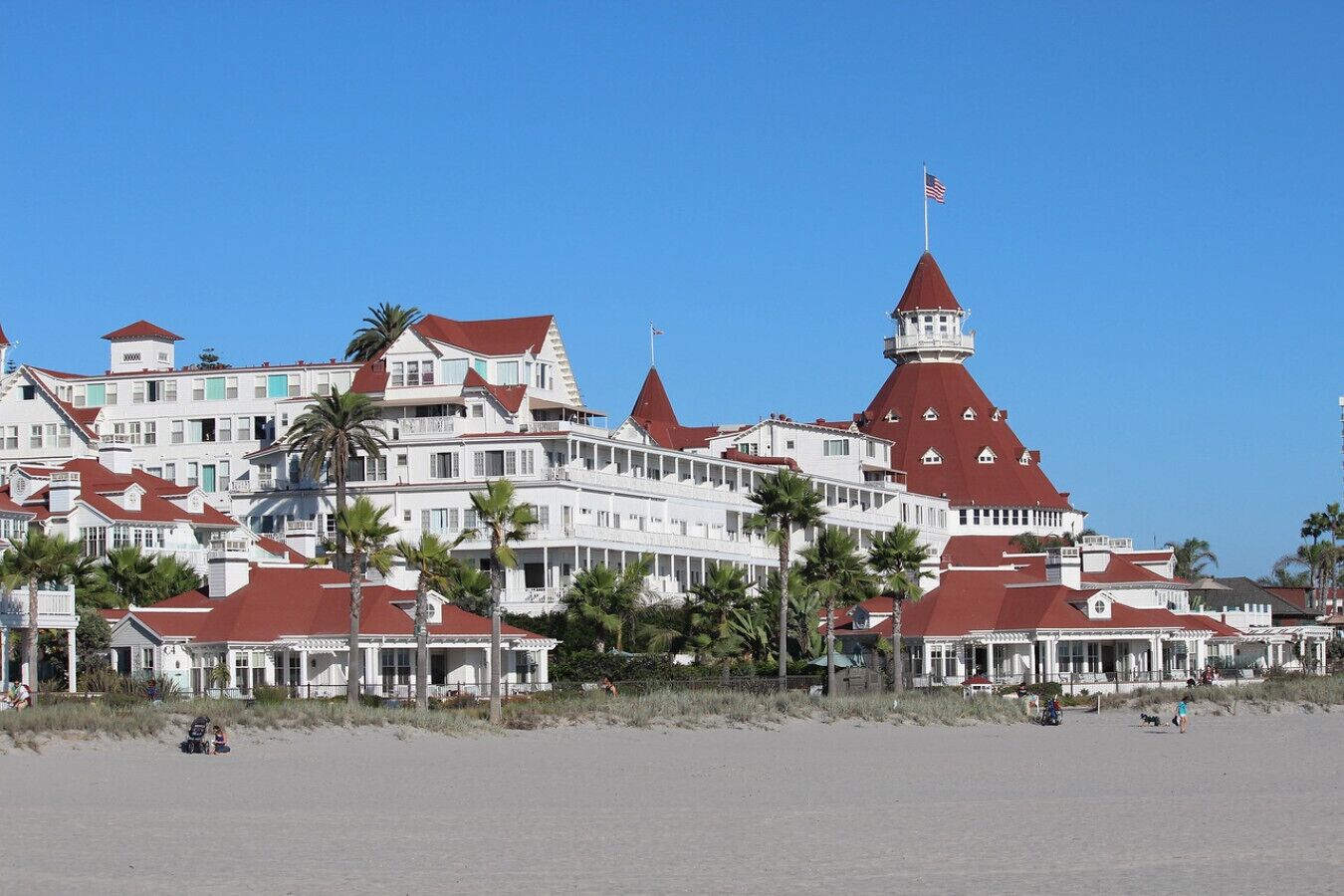 Ett stort hotell med flera våningar, röda tak och vita väggar, som ligger på en strand med palmer.