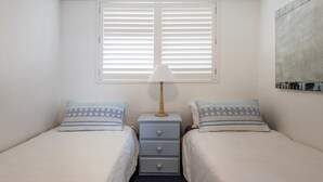 A bedroom with two beds, a bedside table with a lamp, and a window with blinds. - Craigmore On The Beach Unit 11 - Lifestyle Yamba (YAMBA)