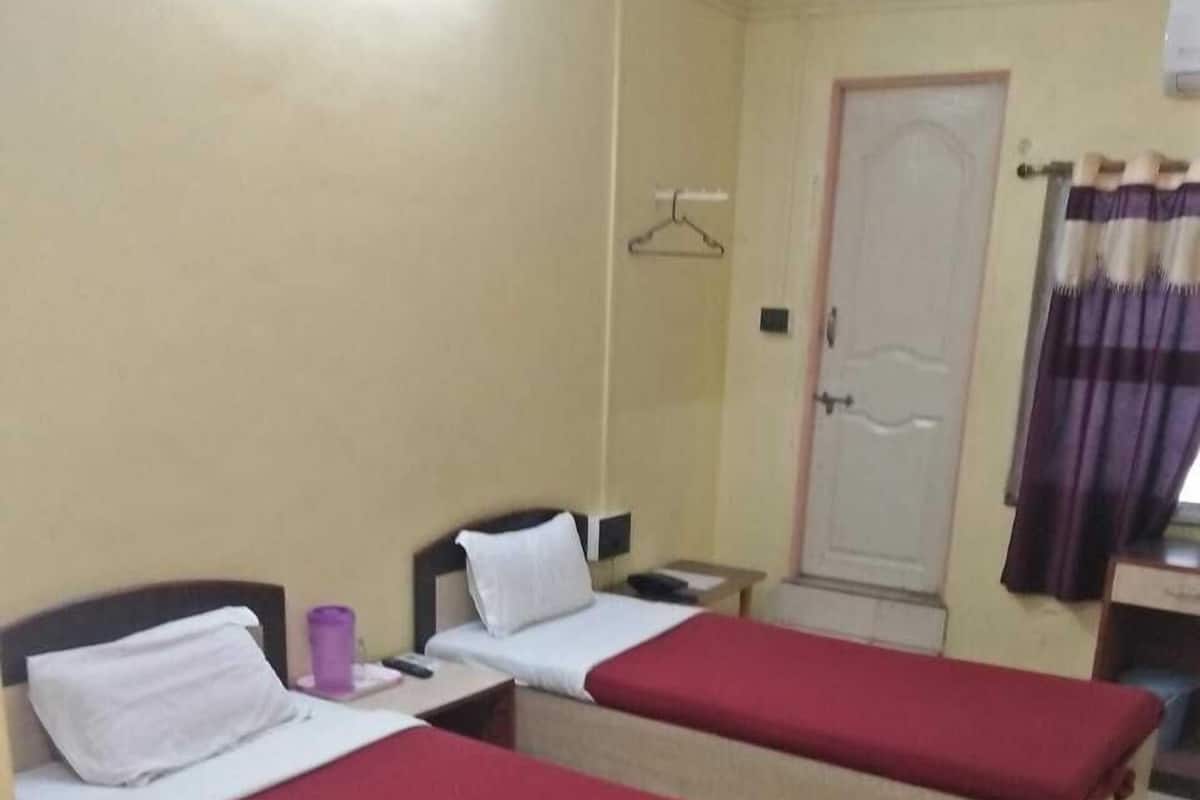 Ein Hotelzimmer mit zwei Einzelbetten, jeweils mit roter Bettdecke und weißen Kissen. Es gibt einen kleinen Tisch mit einer violetten Tasse, eine weiße Tür und ein Fenster mit violetten Vorhängen.
