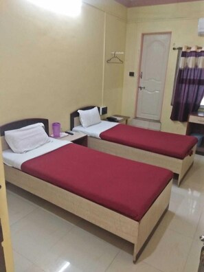 A hotel room with two single beds, each with a red bedspread and white pillows. There is a small table with a purple cup, a white door, and a window with purple curtains. - Hotel Sagar Plaza (Chhatrapati Sambhajinagar)