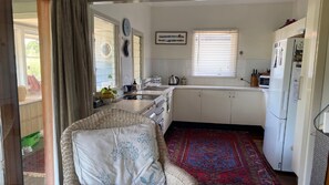 Una cocina compacta con armarios blancos, un lavamanos, una estufa y un refrigerador. Hay una silla de mimbre y una alfombra estampada en la habitación.