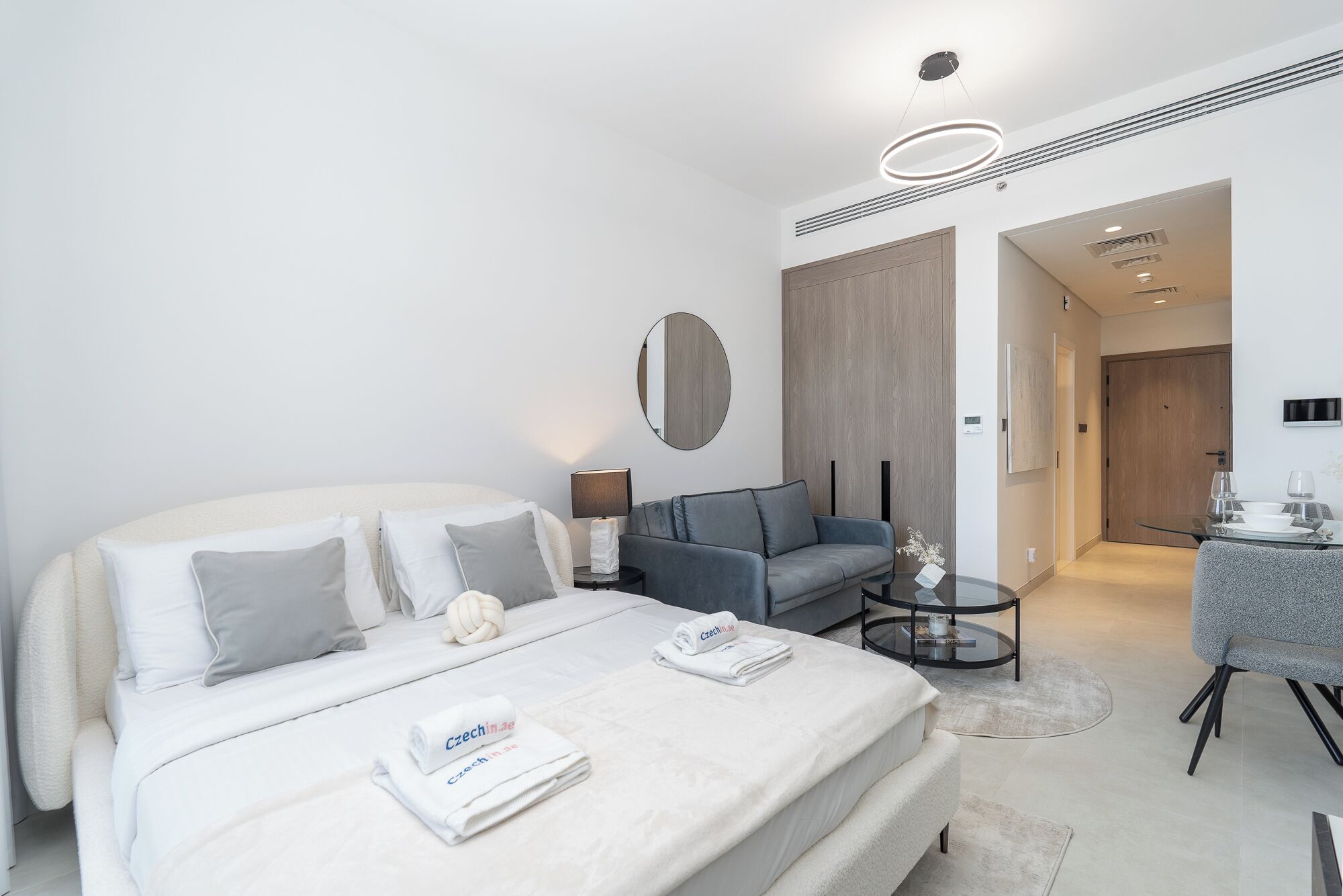 A modern hotel room with a large bed, a gray sofa, a small round table, and a dining area with a table set for two.