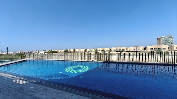 A swimming pool with a clear blue water surface, surrounded by a metal fence and a row of buildings in the background. - TOP Location | Work and Chill near Miracle Garden (Dubai)