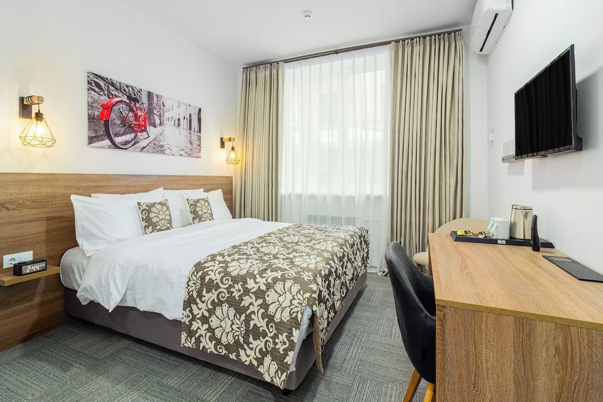 A hotel room with a bed, a desk, a chair, a TV, and a painting on the wall.