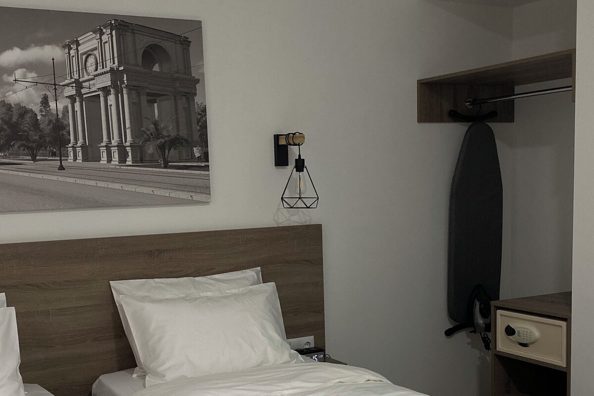 Camera d'albergo con due letti, una testiera in legno, una sveglia a parete, un quadro, una scrivania con uno specchio e una lampada a sospensione.