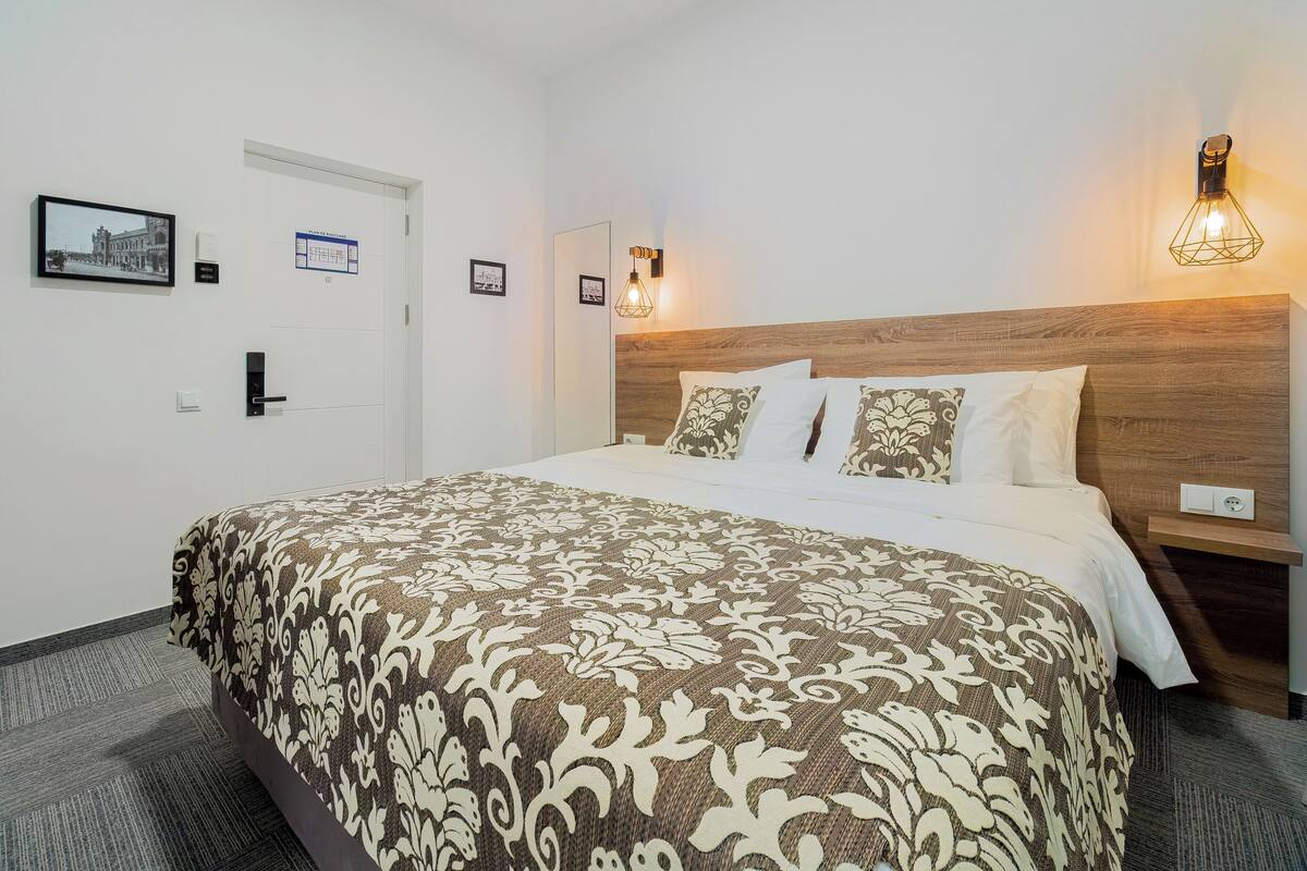 A hotel room with a large bed, a wooden headboard, two bedside tables, a wall-mounted light fixture, and a door with a handle.